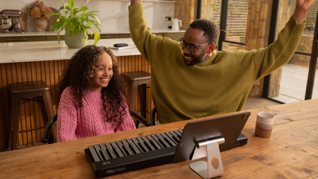 A smiling girl and an enthusiastic man sit at a wooden table in a bright home setting, with a ROLI Piano and a tablet on a stand in front of them.