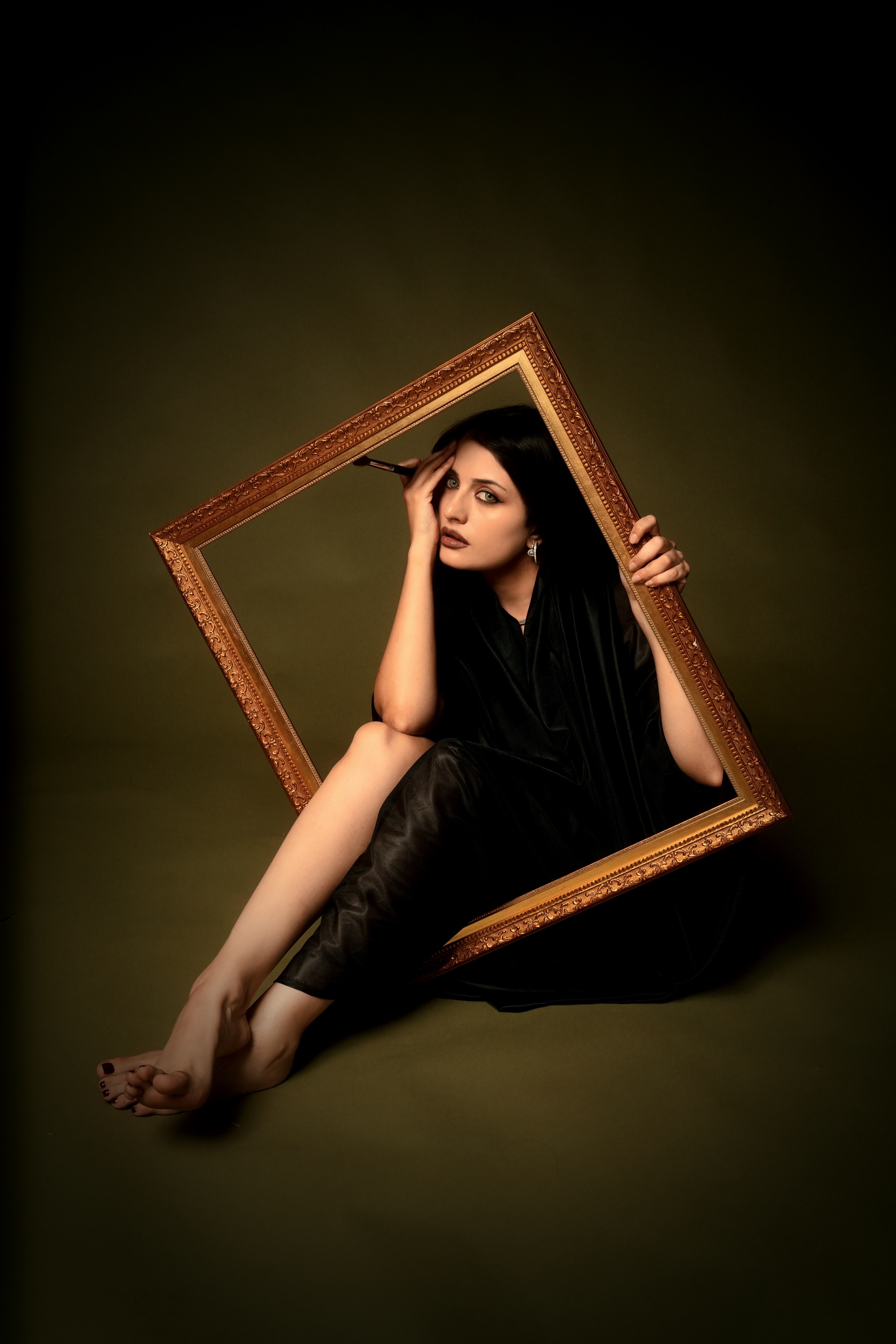 A woman in a black dress sits on the floor holding an ornate empty picture frame, with one leg and arm extending outside the frame and her other hand holding a makeup brush to her face, against a dark olive background.