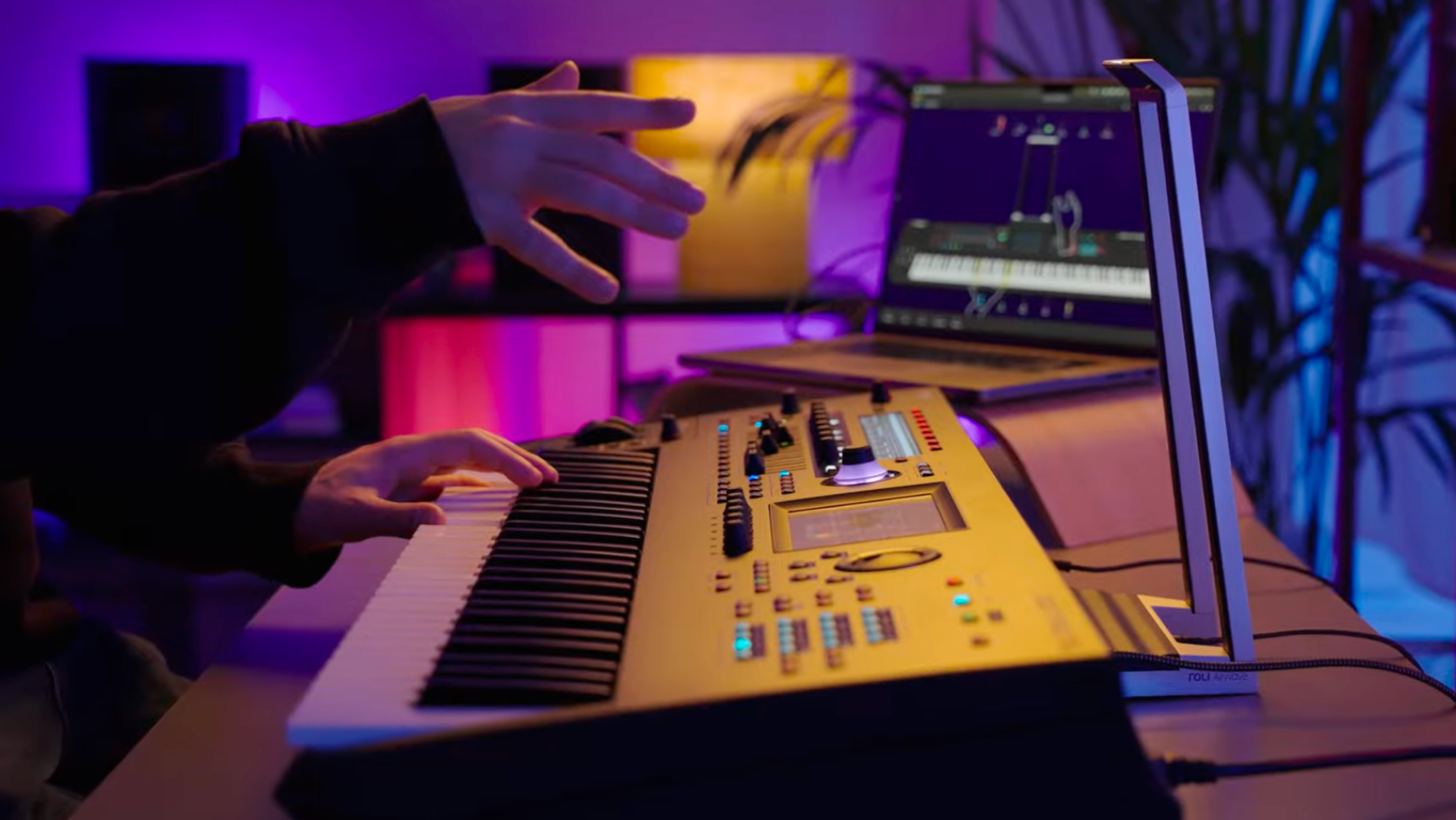 A person playing a Yamaha Montage synthesizer in a home studio, with a ROLI Audio device on the desk and a computer displaying a virtual piano interface in the background, illuminated by colorful ambient lighting.